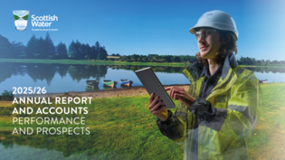Scottish Water male employee wearing hi viz and hat holding an ipad against a backdrop of a reservoir with three boats at the water's edge on a sunny day.