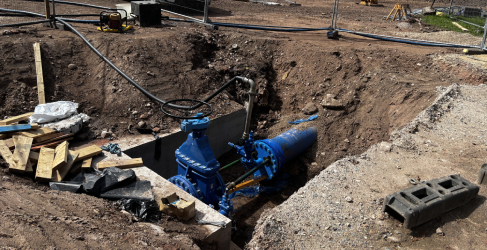A large blue water pipe valve installed in a deep excavation trench at a construction site, surrounded by dirt, gravel, and construction materials.
