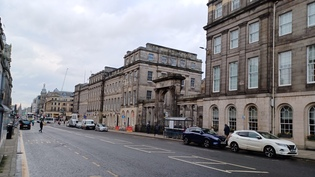 A street with cars parked either side and buildings line the street either side.