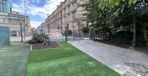 Newly laid astoturf and pavement within a fenced area with buildings in the distance