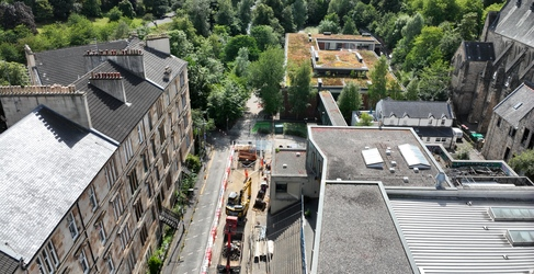 A high angle view of a street with construction work. The street is lined with buildings and there are lots of green trees in the background