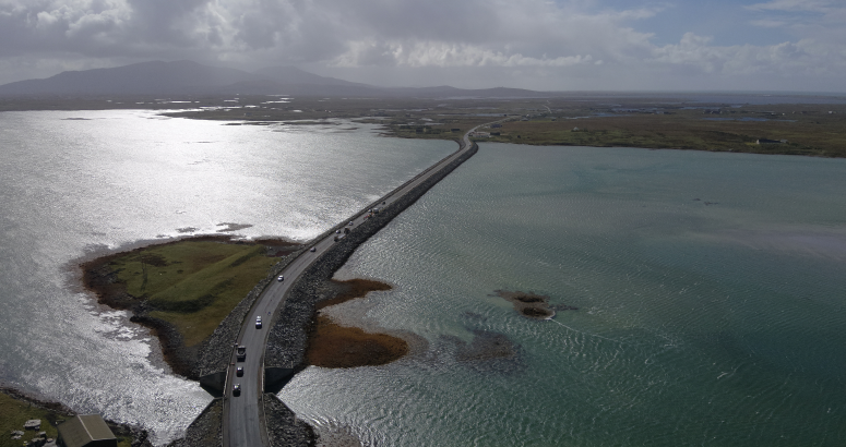 Aerial view of a road curving through a coastal landscape with sunlit water and distant mountains.
