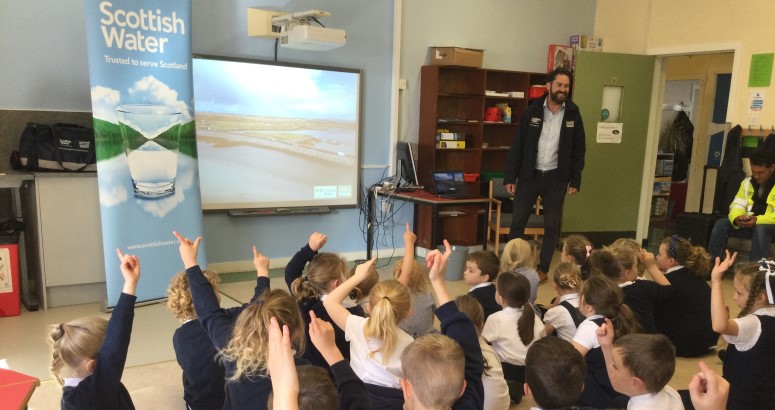 A group of schoolchildren sitting on the floor, raising their hands towards an adult presenter near a screen displaying "Scottish Water" logo and landscape. The setting is an educational classroom.
