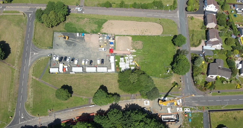 An aerial shot of a construction site compound in a grassy area. A crane is shown lifting equipment into place