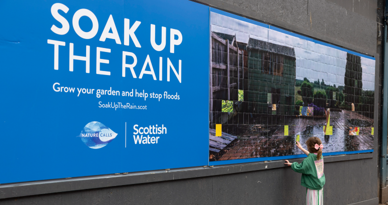 Large billboard on a city street promoting Scottish Water’s “Soak Up the Rain” campaign, featuring a photo of rainwater flooding a garden while a child reaches toward the display.