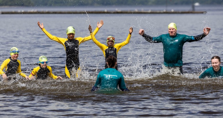 A group of swimmers in the water at Loch Lomond with wet suits and swim caps., 3 are lying in water while 3 are standing with their hands in the air. 