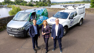 three people standing in front of two branded vehicles with Milngavie Reservoir in the background to announce the new digital partners for Scottish Water