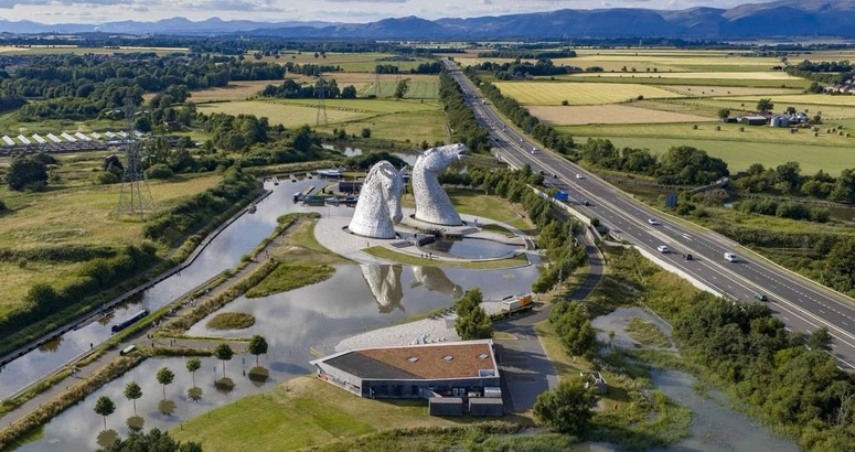 Image shows aerial view of a burst water main with water surrounding The Helix and the Kelpies in Falkirk and a motorway running alongside with countryside and mountain backdrop