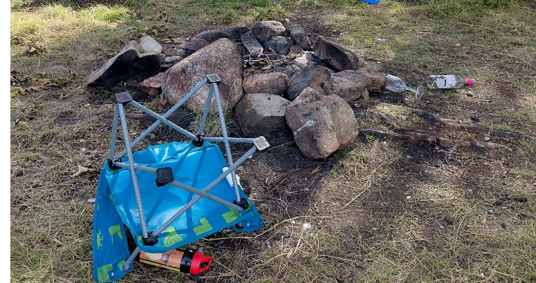 Used campfire left with other camping items such as a knocked over blue chair and bottle of alcohol surrounding it.