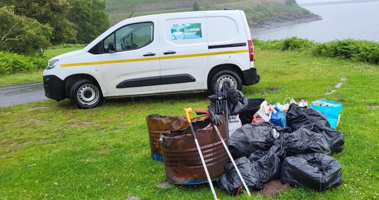 Scottish Water van parked in front of reservoir with pile of rubbish bags and various litter items on grass in front.