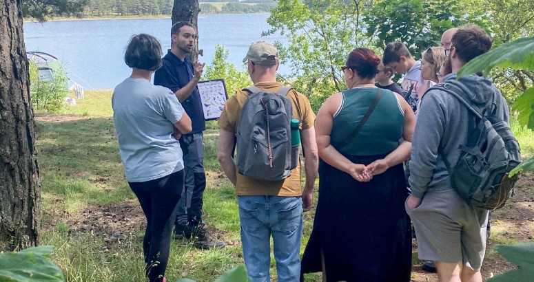 Ranger Josh sharing information with a group of visitors in the woodland at Gladhouse with reservoir in background.