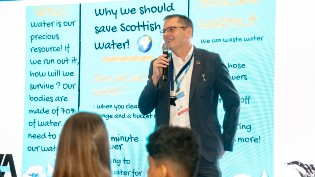 Scottish Water Chief Executive, Alex Plant, presents to primary school kids with microphone and educational blue screen behind him with key SW messages.