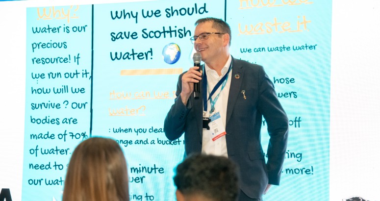 Scottish Water Chief Executive, Alex Plant, presents to primary school kids with microphone and educational blue screen behind him with key SW messages.