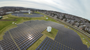 An image taken from a drone which shows a circular formation of PV panels on a grassed area, with houses in the background