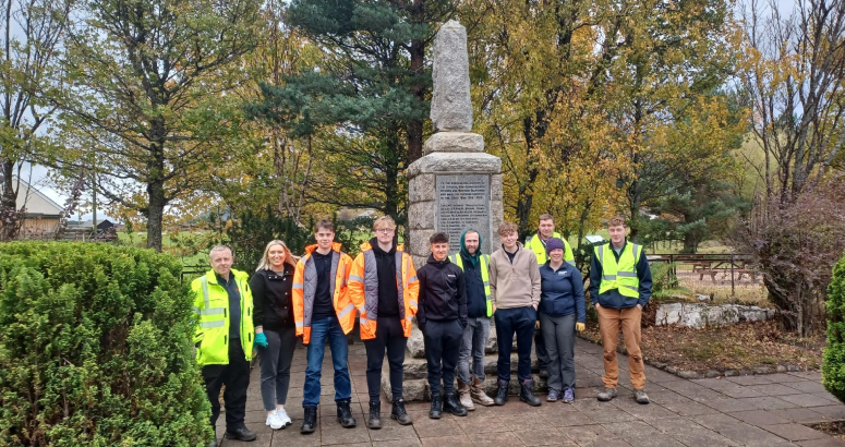 A group of people wearing hi-vis jackets are standing on concrete ground in front of a tall war memorial, with a background of trees in autumnal colours.