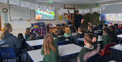 Students in green uniforms sit at tables in a classroom, watching a presentation by an adult at the front. The room is decorated with hanging papers and educational materials.
