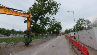A grey road runs down the centre of the image. To the right is orange plastic fencing, and to the left is a yellow piece of heavy plant machinery with trees in the background.