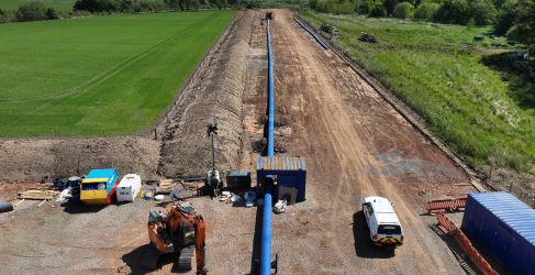 Aerial view of a rural pipeline construction site with machinery, including an excavator and various vehicles, alongside materials laid out on cleared land under a clear sky.