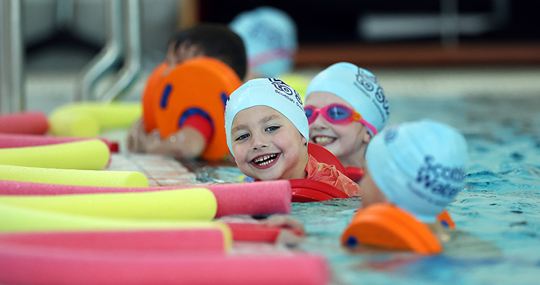 children in swimming pool with smiling face of two, using floats and wearing swim caps