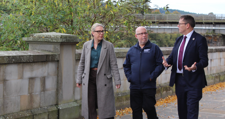 A woman in a long coat is pictured left of the image, with two gentlemen in the centre and right of the picture. They're walking down a pavement with stone wall and trees in the background.