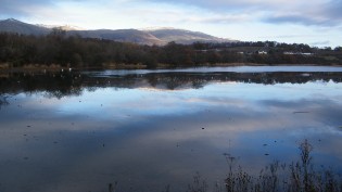 Scenic view of a calm reservoir reflecting the sky, with mountains in the background and sparse vegetation along the shore.